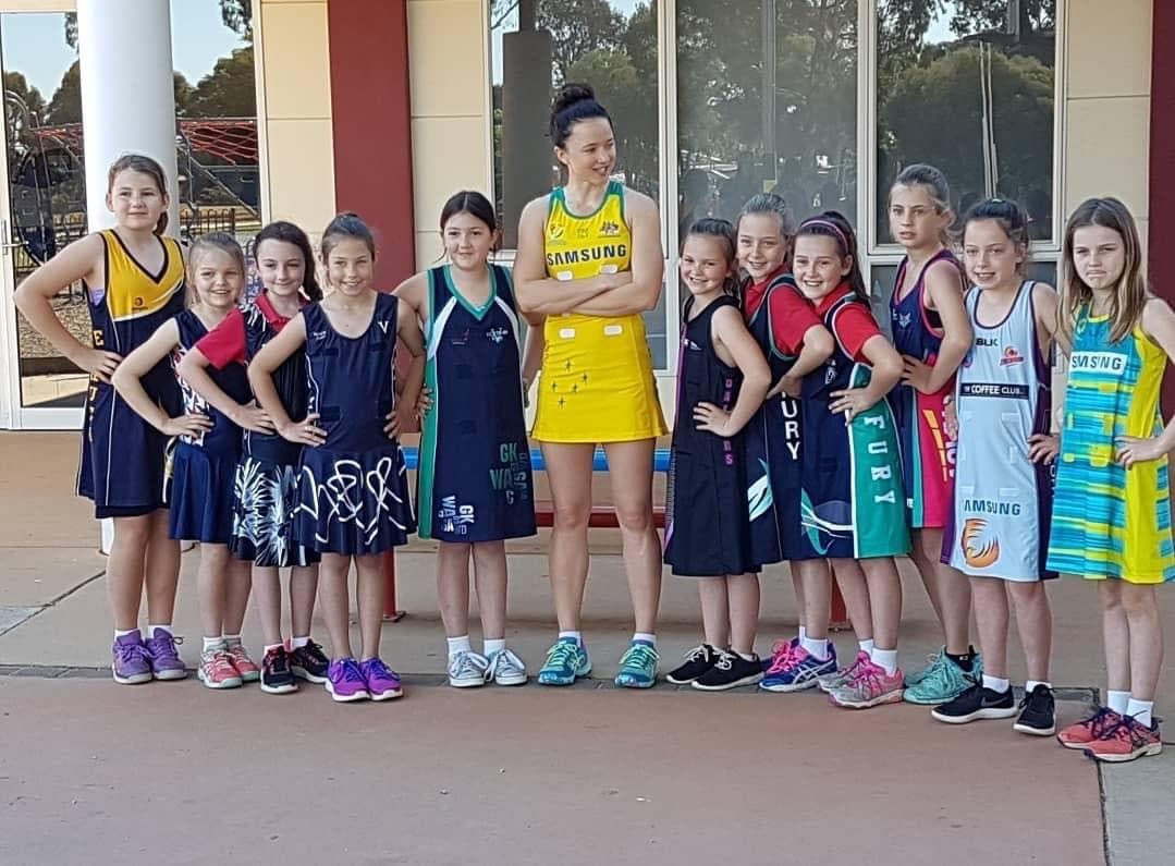 Caitlyn Strachan in the Diamonds gold standing with a line of young players in their club netball dresses, all in matching power poses.